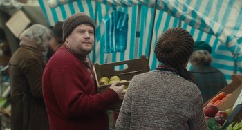 Movie still from “The Lady in the Van” (2015), directed by Nicholas Hytner – A man holding a box of apples while standing next to a woman; Medium shot, Over the shoulder angle
