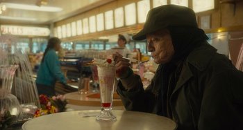 Movie still from “The Lady in the Van” (2015), directed by Nicholas Hytner – An old man sitting at a table with a drink in front of him; Medium shot, Over the shoulder angle
