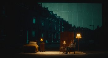 Movie still from “The Lady in the Van” (2015), directed by Nicholas Hytner – A man sitting on a chair in a room with a bed; Wide shot, High angle