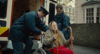 Movie still from “The Lady in the Van” (2015), directed by Nicholas Hytner – An old woman in a wheel chair being assisted by two men; Medium shot, Over the shoulder angle