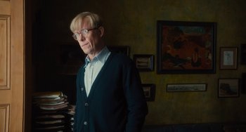 Movie still from “The Lady in the Van” (2015), directed by Nicholas Hytner – A man standing in a room with a bunch of books on the wall behind him; Medium shot, Over the shoulder angle