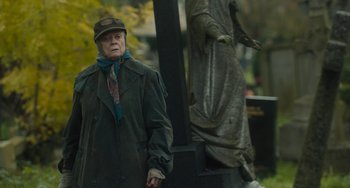 Movie still from “The Lady in the Van” (2015), directed by Nicholas Hytner – An old man in a green coat and a hat standing in front of a statue; Medium shot, Over the shoulder angle