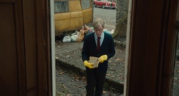 Movie still from “The Lady in the Van” (2015), directed by Nicholas Hytner – An older man wearing yellow rubber gloves while reading a book; Medium shot, Low angle