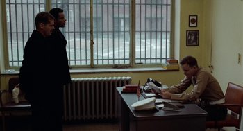 Movie still from “The Last Detail” (1973), directed by Hal Ashby – A man sitting at a desk in front of another man; Wide shot, Over the shoulder angle