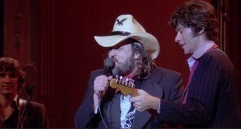 Movie still from “The Last Waltz” (1978), directed by Martin Scorsese – A man wearing a cowboy hat holding a microphone and a guitar; Medium shot, Over the shoulder angle