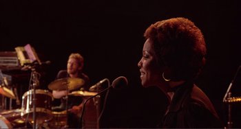 Movie still from “The Last Waltz” (1978), directed by Martin Scorsese – A woman standing at a microphone in front of a man playing an instrument; Medium shot, Over the shoulder angle