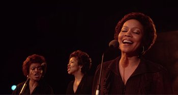 Movie still from “The Last Waltz” (1978), directed by Martin Scorsese – A group of women singing into microphones in front of a black background; Medium shot, Low angle