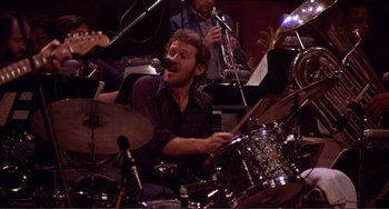 Movie still from “The Last Waltz” (1978), directed by Martin Scorsese – A man playing drums in front of a group of other musicians; Close Up shot, High angle