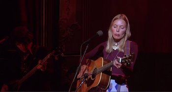 Movie still from “The Last Waltz” (1978), directed by Martin Scorsese – A woman is playing an acoustic guitar in front of a microphone; Medium shot, Low angle
