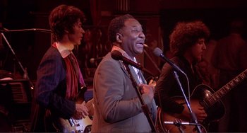 Movie still from “The Last Waltz” (1978), directed by Martin Scorsese – A group of people that are standing around a microphone; Medium shot, Low angle