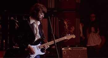 Movie still from “The Last Waltz” (1978), directed by Martin Scorsese – A man with a beard playing an electric guitar in front of a microphone; Medium shot, Low angle