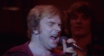 Movie still from “The Last Waltz” (1978), directed by Martin Scorsese – A man is singing into a microphone in front of a group of people; Close Up shot, Low angle