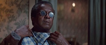 Movie still from “The Learning Tree” (1969), directed by Gordon Parks – An older man wearing round sunglasses and a neck tie adjusts his neck tie; Close Up shot, Over the shoulder angle