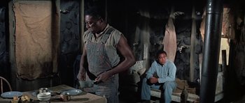 Movie still from “The Learning Tree” (1969), directed by Gordon Parks – A man standing next to a man sitting on a bench; Medium shot, Low angle