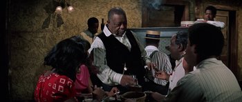 Movie still from “The Learning Tree” (1969), directed by Gordon Parks – An older man lighting a cigarette at a table; Medium shot, Over the shoulder angle