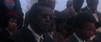 Movie still from “The Learning Tree” (1969), directed by Gordon Parks – A man in a suit and round sunglasses is in a crowd of people; Close Up shot, Over the shoulder angle