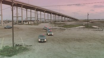 Movie still from “The Legend of Billie Jean” (1985), directed by Matthew Robbins – Three police cars are parked in a parking lot near a bridge; Extreme Wide shot, High angle
