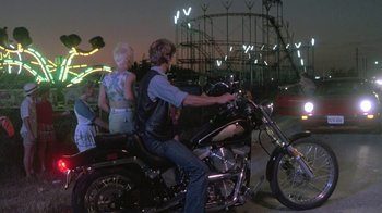 Movie still from “The Legend of Billie Jean” (1985), directed by Matthew Robbins – A man on a motorcycle with a woman on a roller coaster in the background; Wide shot, Over the shoulder angle