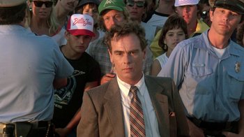 Movie still from “The Legend of Billie Jean” (1985), directed by Matthew Robbins – A man in a suit standing in front of a group of people; Close Up shot, Over the shoulder angle