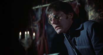 Movie still from “The Legend of Hell House” (1973), directed by John Hough – A man with glasses is sitting in a dark room; Close Up shot, Low angle