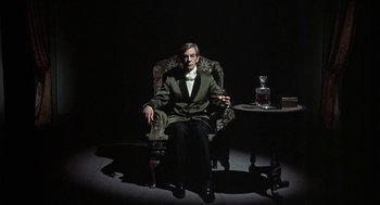 Movie still from “The Legend of Hell House” (1973), directed by John Hough – A man sitting in a chair with a cigarette in his mouth; Medium shot, Low angle