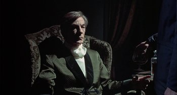 Movie still from “The Legend of Hell House” (1973), directed by John Hough – An older man sitting in a chair wearing a suit and tie; Medium shot, Low angle