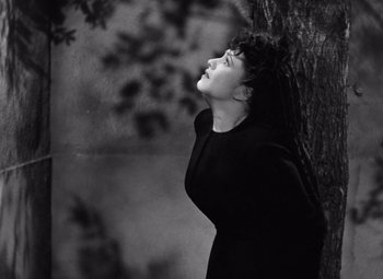Movie still from “The Leopard Man” (1943), directed by Jacques Tourneur – A black and white photo of a woman leaning up against a tree; Medium shot, High angle