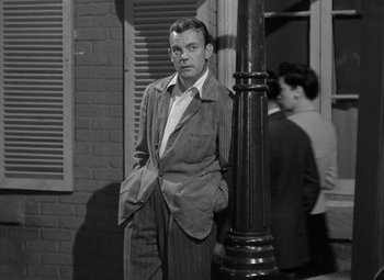 Movie still from “The Leopard Man” (1943), directed by Jacques Tourneur – A black and white photo of a man leaning against a pole; Medium shot, Low angle