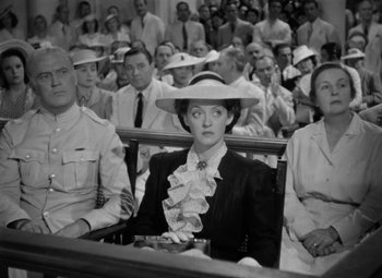 Movie still from “The Letter” (1940), directed by William Wyler – A woman in a hat sitting in front of a group of people; Medium shot, High angle