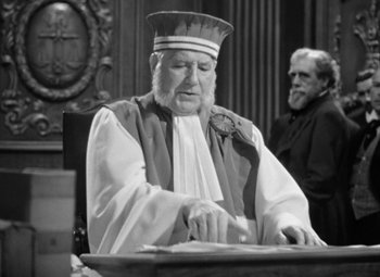 Movie still from “The Life of Emile Zola” (1937), directed by William Dieterle – An older man in a priest's outfit sitting at a table; Medium shot, Low angle