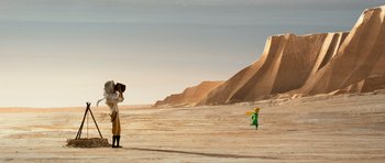 Two people in a desert area with a green figure in front of a mountain