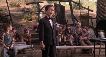 Movie still from “The Little Rascals” (1994), directed by Penelope Spheeris – A man in a suit and bow tie standing in front of a group of people; Medium shot, Over the shoulder angle