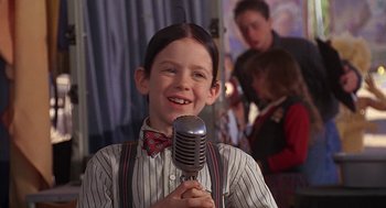Movie still from “The Little Rascals” (1994), directed by Penelope Spheeris – A young boy holding a microphone in front of a group of people; Close Up shot, Over the shoulder angle