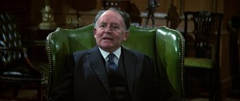 Movie still from “The Living Daylights” (1987), directed by John Glen – An older man wearing a suit and tie sitting in a chair; Close Up shot, High angle