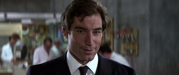 Movie still from “The Living Daylights” (1987), directed by John Glen – A man in a suit and tie smiling for the camera; Close Up shot, Over the shoulder angle