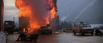 Movie still from “The Living Daylights” (1987), directed by John Glen – A car on fire in the middle of a road; Wide shot, High angle