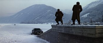 Movie still from “The Living Daylights” (1987), directed by John Glen – Two men standing on the side of a wall near a parked car; Wide shot, High angle