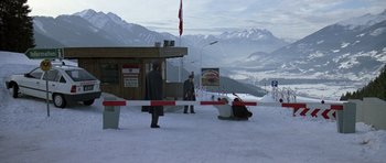 Movie still from “The Living Daylights” (1987), directed by John Glen – A man laying in the snow next to a red and white fence; Extreme Wide shot, High angle