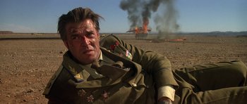 Movie still from “The Living Daylights” (1987), directed by John Glen – A man in a military uniform laying on the ground in front of a burning vehicle; Close Up shot, Low angle