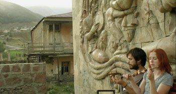 Movie still from “The Loneliest Planet” (2011), directed by Julia Loktev – A man playing a flute in front of a wall with carvings on it; Wide shot, Low angle