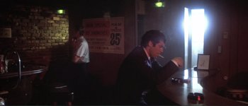 Movie still from “The Long Goodbye” (1973), directed by Robert Altman – A man sitting at a table smoking a cigarette in front of a sign; Medium shot, Over the shoulder angle