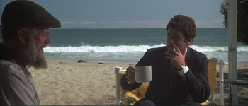Movie still from “The Long Goodbye” (1973), directed by Robert Altman – A man sitting on the beach holding a coffee cup; Medium shot, High angle