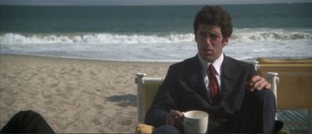 Movie still from “The Long Goodbye” (1973), directed by Robert Altman – A man in a suit and tie sitting on the beach drinking coffee; Medium shot, Low angle