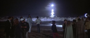 Movie still from “The Long Goodbye” (1973), directed by Robert Altman – A group of people standing on top of a sandy beach at night; Extreme Wide shot, High angle