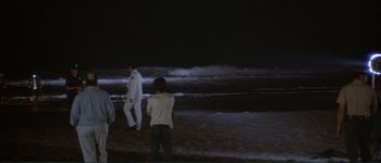 Movie still from “The Long Goodbye” (1973), directed by Robert Altman – A group of people standing on top of a sandy beach at night; Extreme Wide shot, High angle