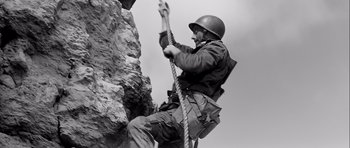Movie still from “The Longest Day” (1962), directed by Ken Annakin – A man in a helmet climbing up a rock wall; Medium shot, Low angle