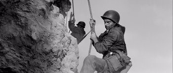 Movie still from “The Longest Day” (1962), directed by Ken Annakin – A man in a helmet is climbing a rock; Medium shot, Low angle