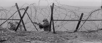 Movie still from “The Longest Day” (1962), directed by Ken Annakin – A man in a raincoat is standing next to barbed wire; Wide shot, Low angle