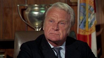 Movie still from “The Longest Yard” (1974), directed by Robert Aldrich – An older man wearing a suit and tie in front of a trophy; Close Up shot, Low angle