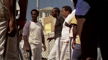 Movie still from “The Longest Yard” (1974), directed by Robert Aldrich – A group of men standing next to each other near a chain link fence; Medium shot, High angle